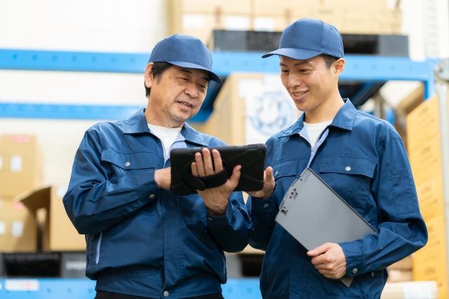 青い作業着を着た二人の男性がタブレット端末を見ながら笑顔を浮かべている